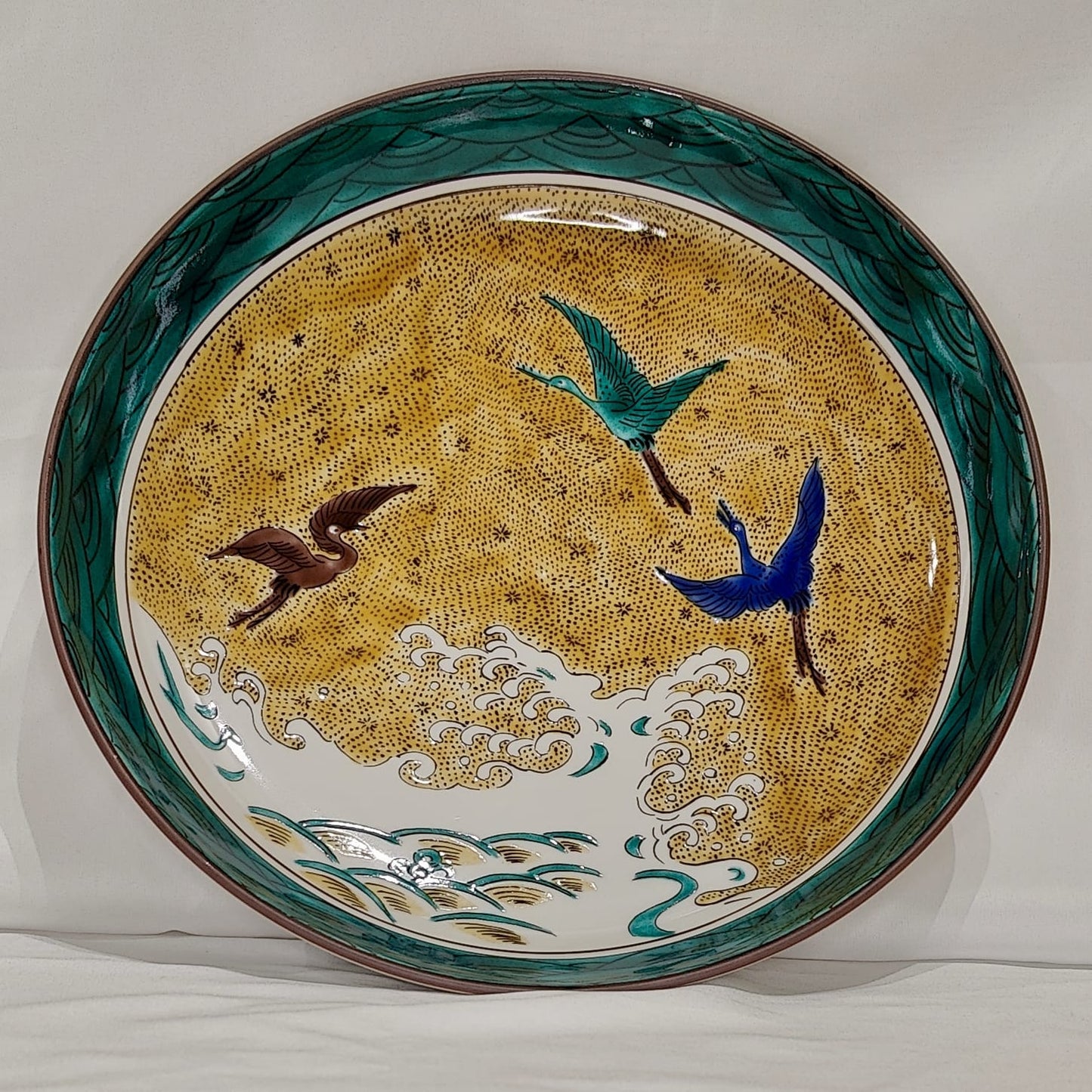 Assiette japonaise décorée de grues en vol et de vagues sur fond doré et vert.